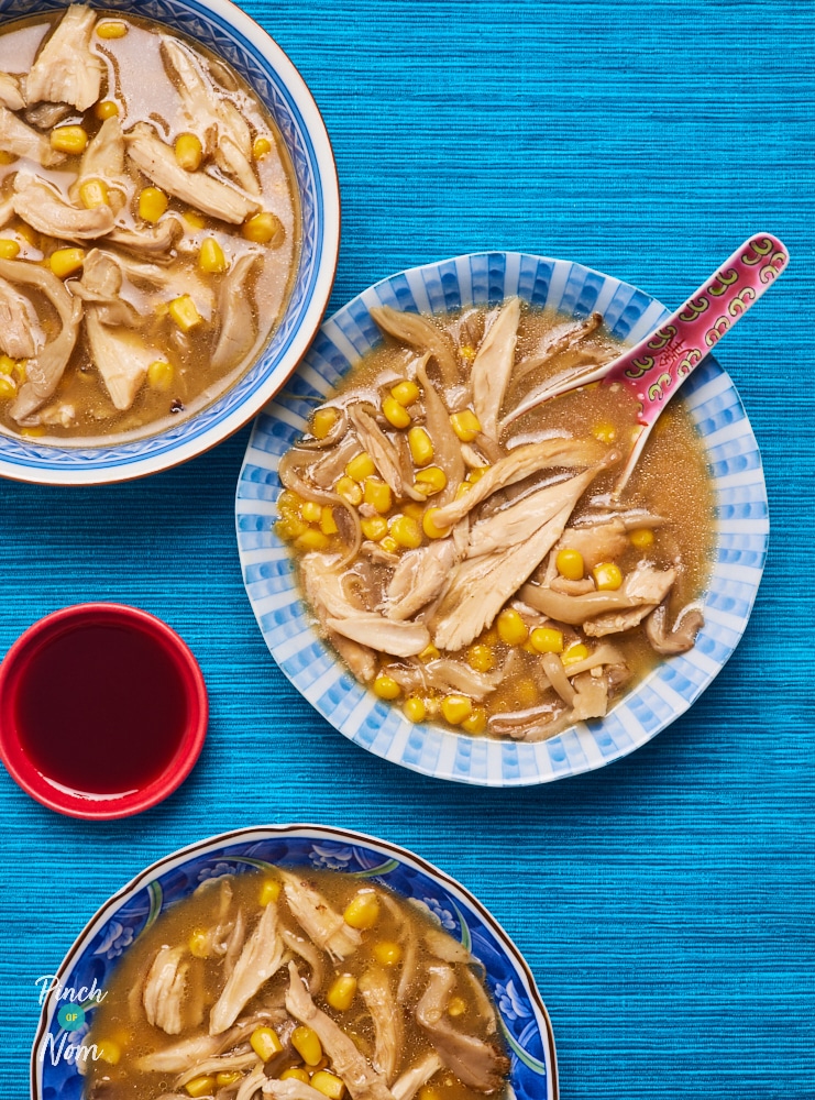 Three servings of Pinch of Nom's Chicken, Mushroom and Sweetcorn Soup are served in blue bowls, set on a bright blue surface. An Asian-style spoon rests in the central bowl, and a small red bowl of soy sauce is on the centre of the table.