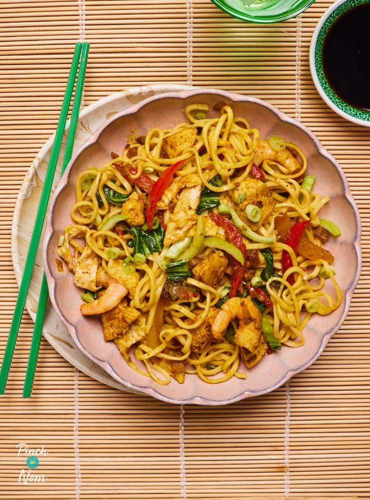 A table is set with a plate of Pinch of Nom's colourful Singapore-Style Noodles; colourful chopped veggies, chicken chunks and prawns are stirred through. Chopsticks and soy sauce wait to the side, ready to tuck in.