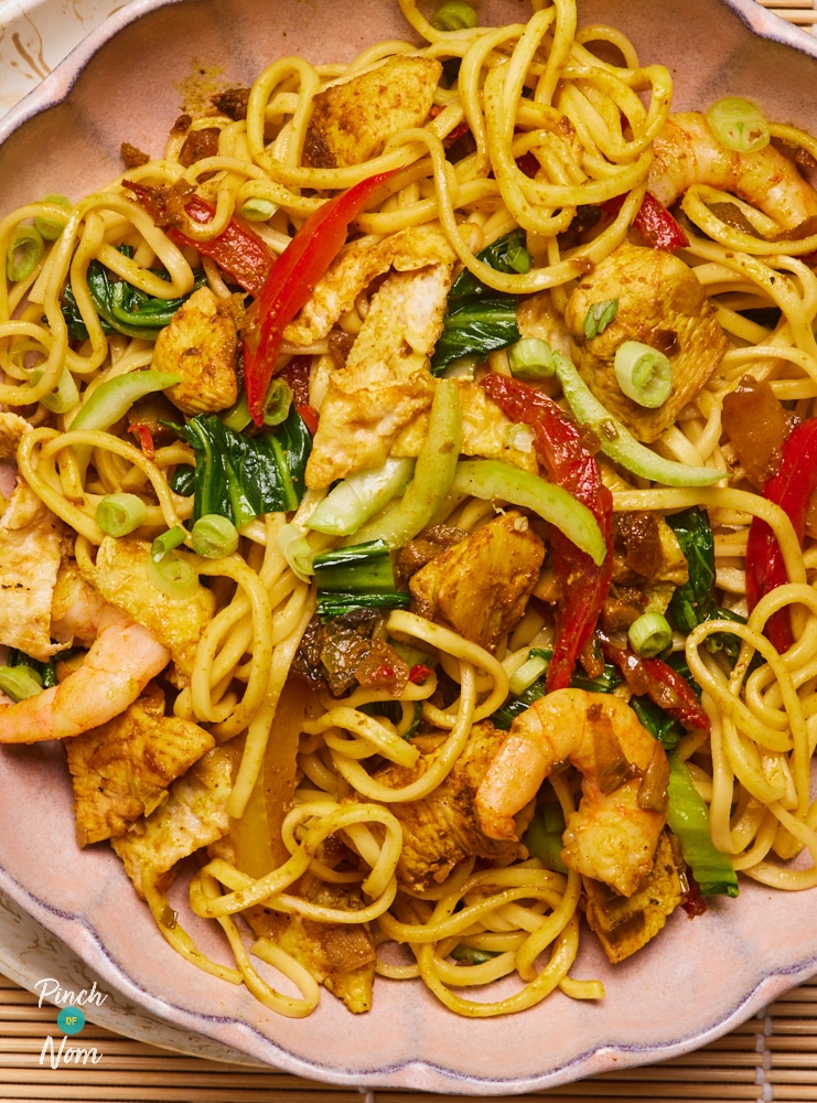 A close-up of Pinch of Nom's Singapore-Style Noodles served on a pink plate; colourful veggies, chicken chunks and prawns can be seen stirred through the noodles.