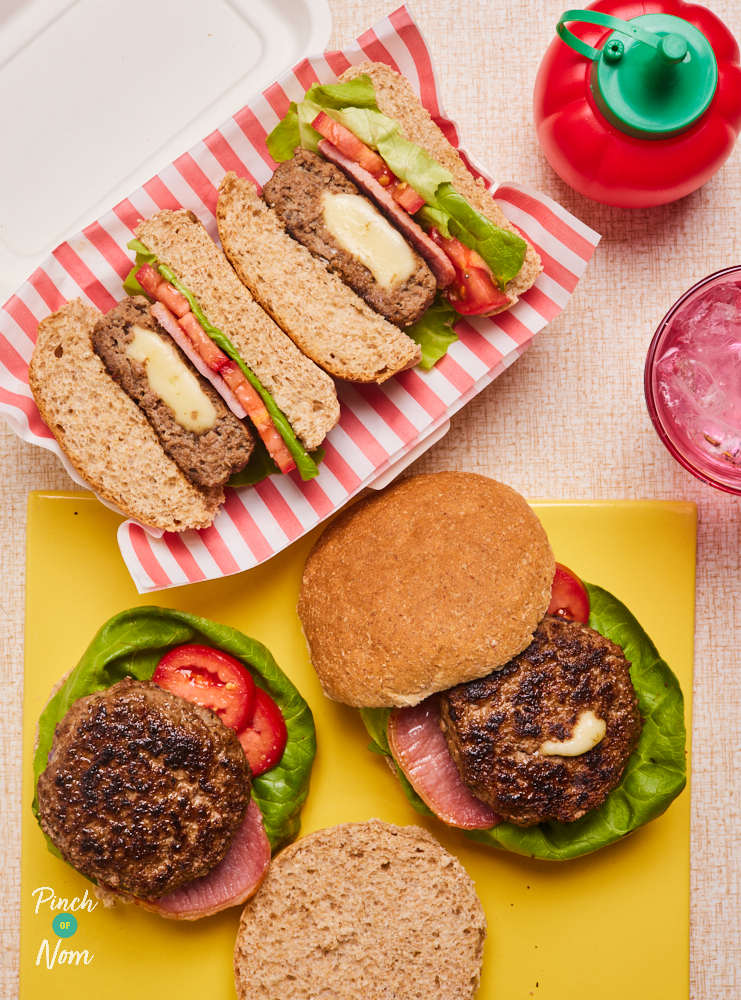 Pinch of Nom's Melt-in-the-Middle Bacon Cheeseburgers are served in a soft bun, with slices of lettuce and tomato. Two of the burgers are partly assembled on a bright yellow chopping board, and another fully-assembled burger has been cut in half and served in a container lined with takeaway-style red and white striped paper. The cross section of the burger can be seen, with melted cheese oozing out of the middle.