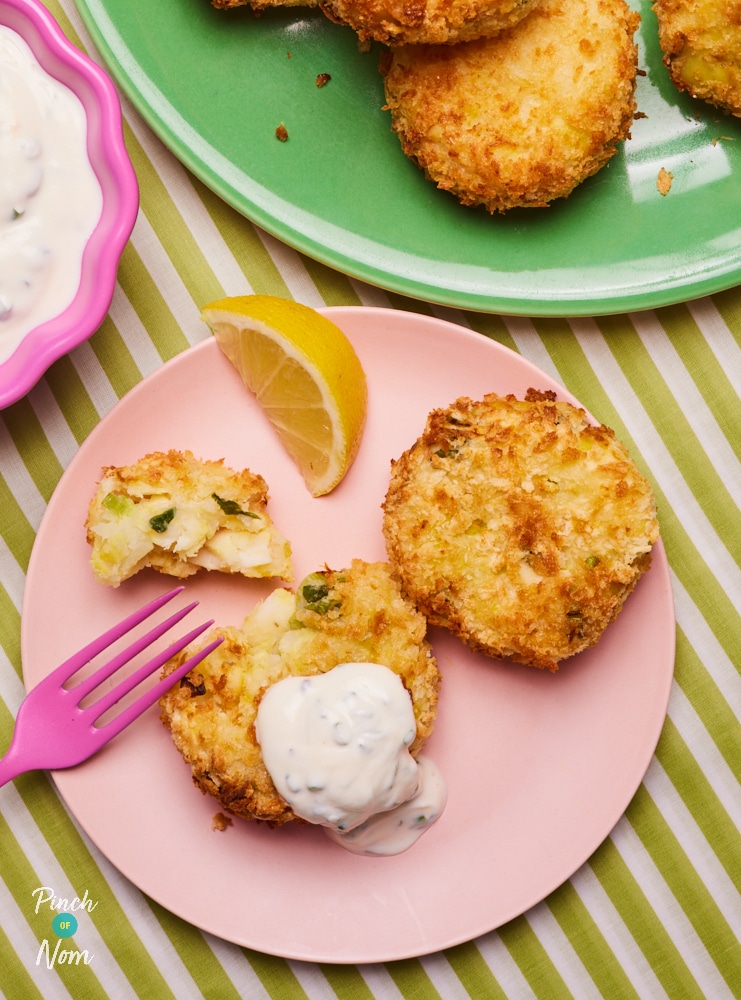 On a small pink plate, a pink fork is cutting into one of Pinch of Nom's slimming-friendly Smoked Haddock and Spring Onion fishcake, with a lemon wedge to one side.