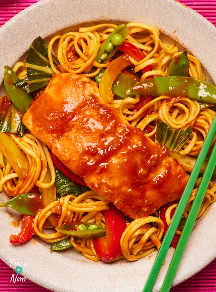 A close-up of a plate of noodles is topped with Pinch of Nom's Teriyaki Chilli Salmon. There are plenty of colourful veggies stirred through the noodles, and chopsticks are set to the side waiting to dig in.