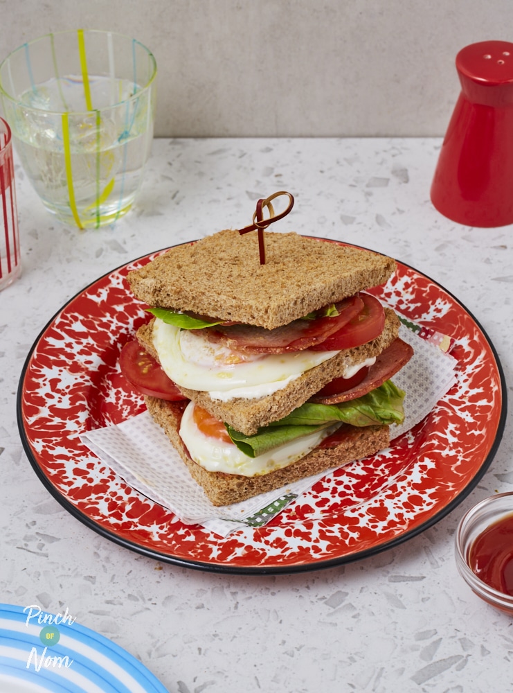 Pinch of Nom's BLT Sandwich is served on a red and white patterned plate. Three slices of bread are stacked up with bacon, fried eggs, fresh tomato, mayonnaise and lettuce.