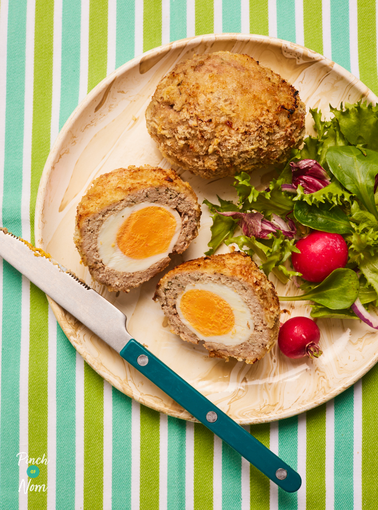 Pinch of Nom's slimming-friendly scotch eggs are served on a pastel pink plate, set on a green and blue striped tablecloth. One of the scotch eggs has been cut in half, revealing the yellow yolk in the centre. A side salad is served with the 2 eggs.