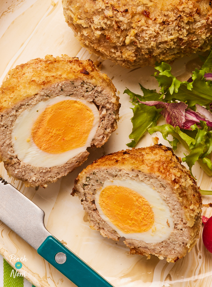 A close-up shows Pinch of Nom's slimming-friendly scotch eggs served on a pastel pink plate. One of the scotch eggs has been cut in half, revealing the yellow yolk in the centre. A side salad is served with the 2 eggs.