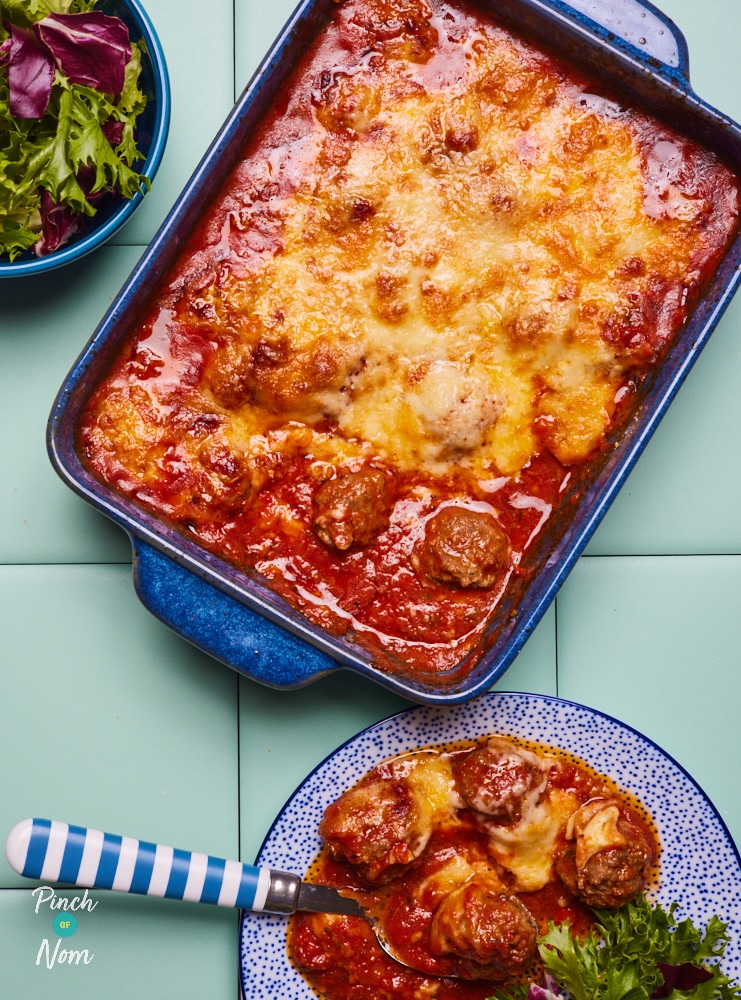 Pinch of Nom's slimming-friendly Baked Spicy Meatballs recipe is served in a large casserole dish, fresh from the oven at the centre of a blue worktop. A serving has been plated up to one side with a fork waiting to tuck in. The bake is topped with a bubbly layer of browned mozzarella and Parmesan.