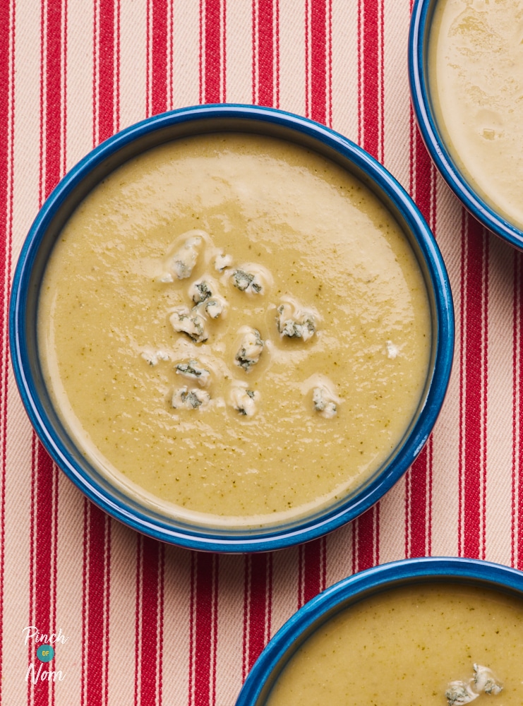 A portion of Pinch of Nom's slimming-friendly Cauliflower and Broccoli soup is served in a bright blue bowl on a pink and red striped tabletop. The portion is topped with crumbled Stilton cheese.