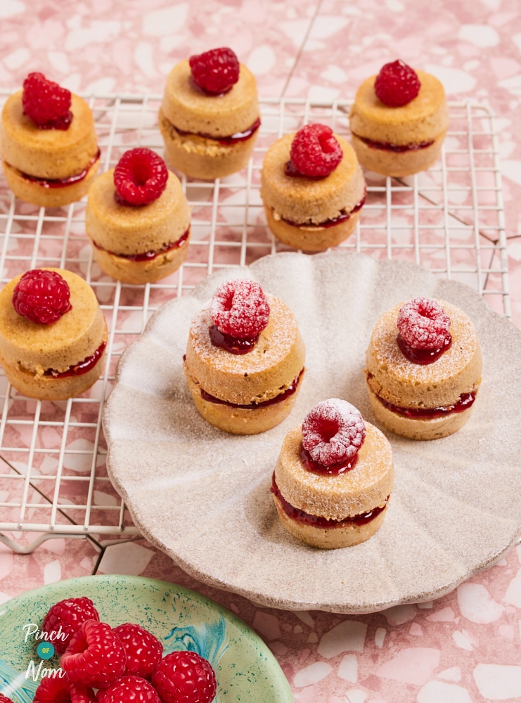 Pinch of Nom's Mini Victoria Sponge Cakes are served topped with fresh raspberries and dusted in icing sugar. Several cakes are resting on a wire cooling rack, and 3 have been arranged on a white scalloped plate. Extra fresh raspberries are on a small green plate nearby.