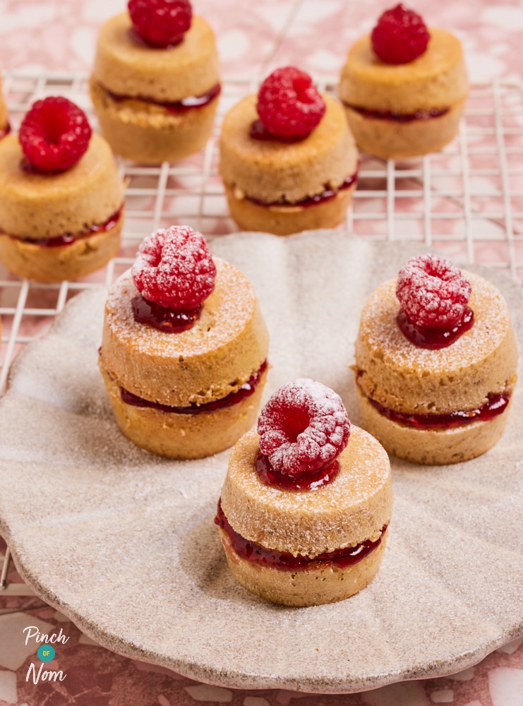 Pinch of Nom's Mini Victoria Sponge Cakes are served topped with fresh raspberries and dusted in icing sugar. Several cakes are resting on a wire cooling rack, and 3 have been arranged on a white scalloped plate.