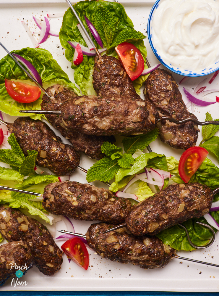 A close-up of Pinch of Nom's Beef Kofta Kebabs on a bed of colourful lettuce leaves, onions and chopped tomatoes. A dipping sauce of fat-free Greek yoghurt is presented to one side. Each kebab is grilled until browned on the outside and cooked through.