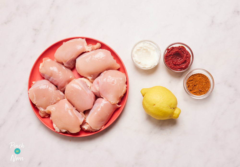 The ingredients for Pinch of Nom's slimming-friendly Cajun-Style Chicken Thighs are laid out on a kitchen work surface. The raw chicken is set out on a red plate, and all the spices are measured into small glass bowls.