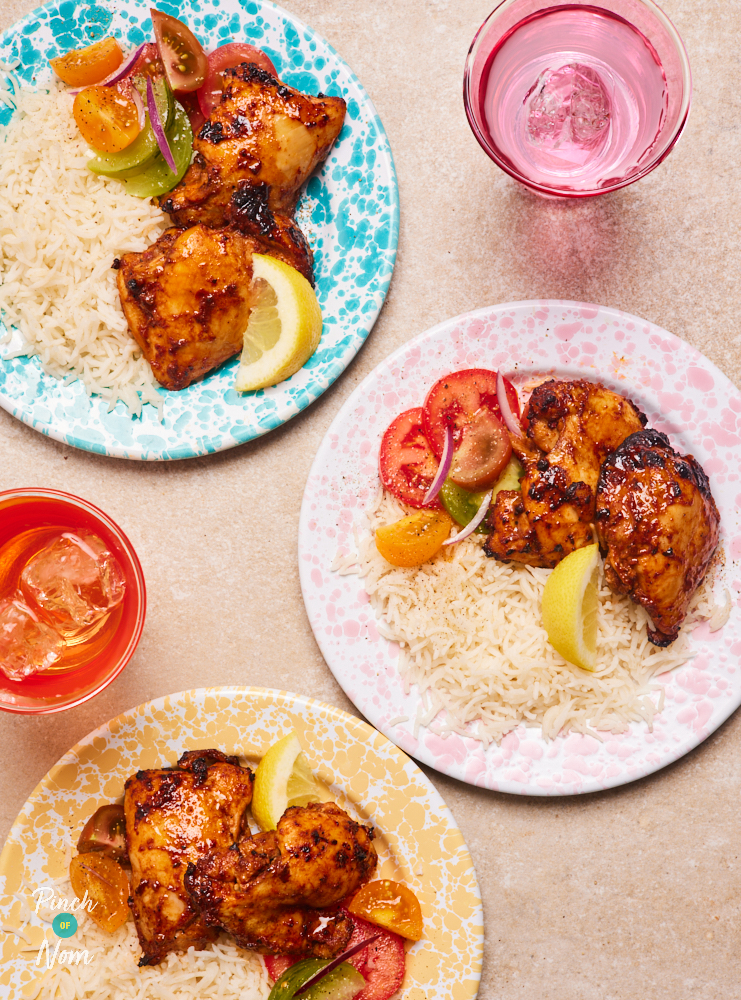 Three portions of Pinch of Nom's Cajun-Style Chicken Thighs are served on pastel-coloured patterned plates. Each portion is served with a spoonful of fluffy rice, vegetables and a wedge of lemon. Glasses of ice water are set on the table, in-between the plates.