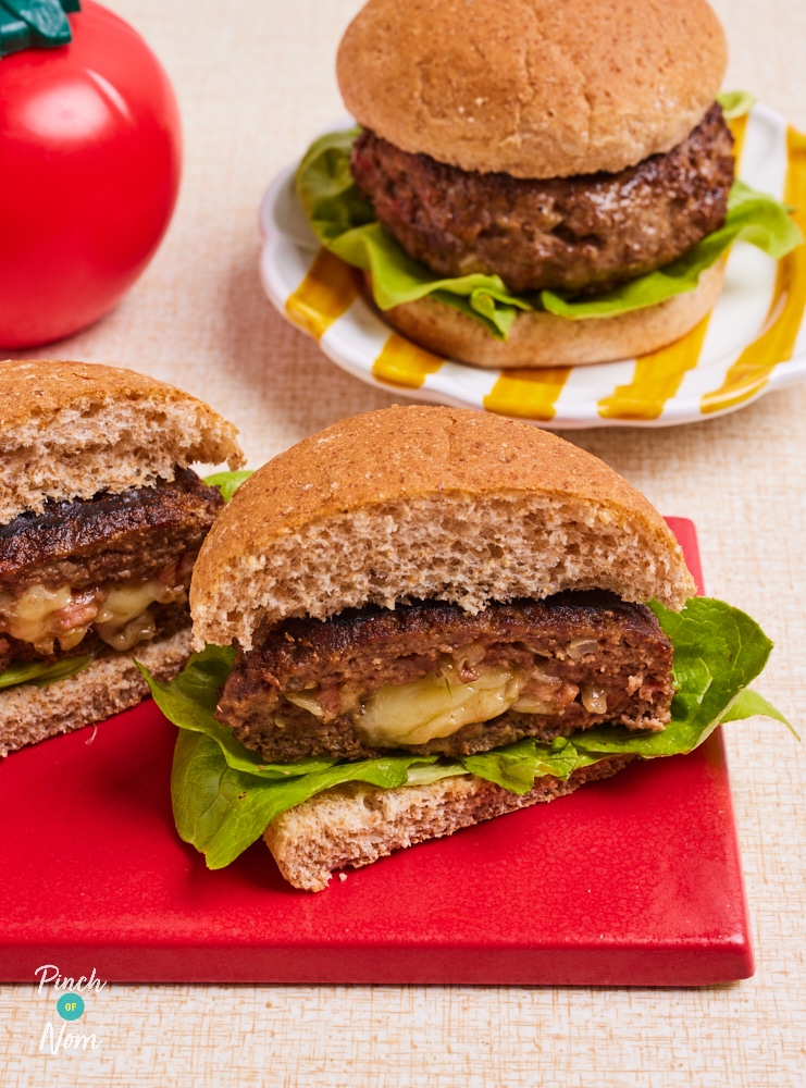 Pinch of Nom's Cheese Stuffed Burgers are served in wholemeal buns, with lettuce leaves. One burger waits on a white and yellow striped plate, the other is served on a red board, cut in half to reveal the oozy cheese centre. A ketchup bottle in the shape of a tomato is in the background.