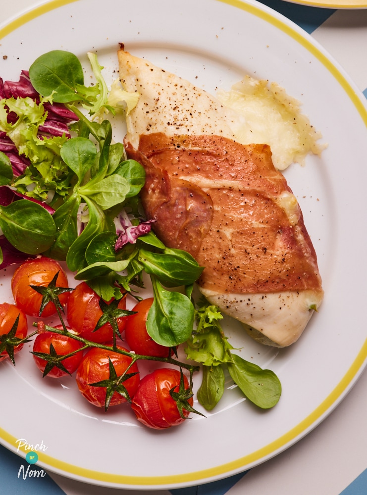 A table is set with a plateful of Pinch of Nom's slimming-friendly Mozzarella and Asparagus Chicken with a side of leafy greens and vine tomatoes.