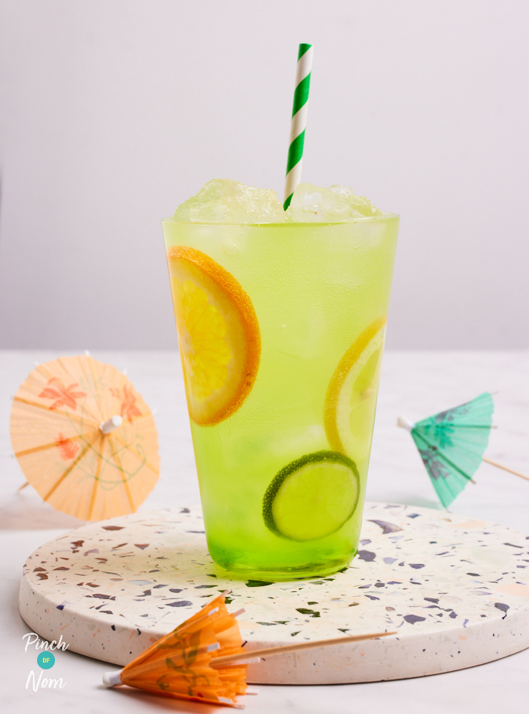 Pinch of Nom's slimming-friendly June Bug Cocktail is served in a tall glass with a green and white striped straw, placed on a speckled ceramic coaster. The cocktail has a vibrant lime green colour, and slices of fresh fruit can be seen inside the glass. Decorative cocktail umbrellas are scattered on the table, around the glass.