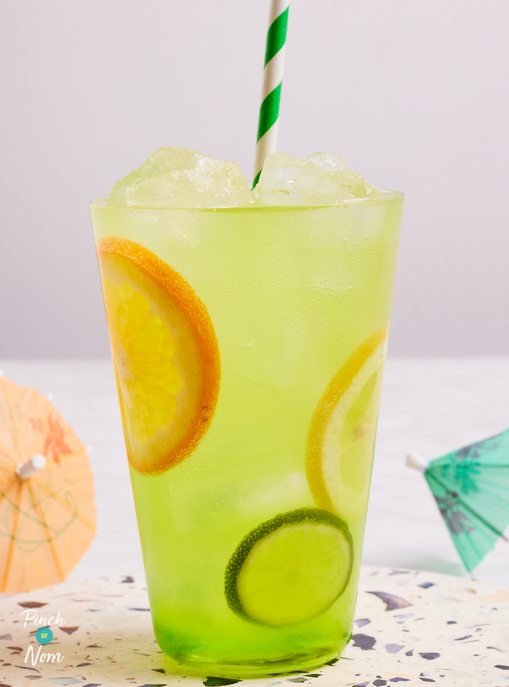 A close up shows Pinch of Nom's slimming-friendly June Bug Cocktail, served in a tall glass with a green and white striped straw, placed on a speckled ceramic coaster. The cocktail has a vibrant lime green colour, and slices of fresh fruit can be seen inside the glass.