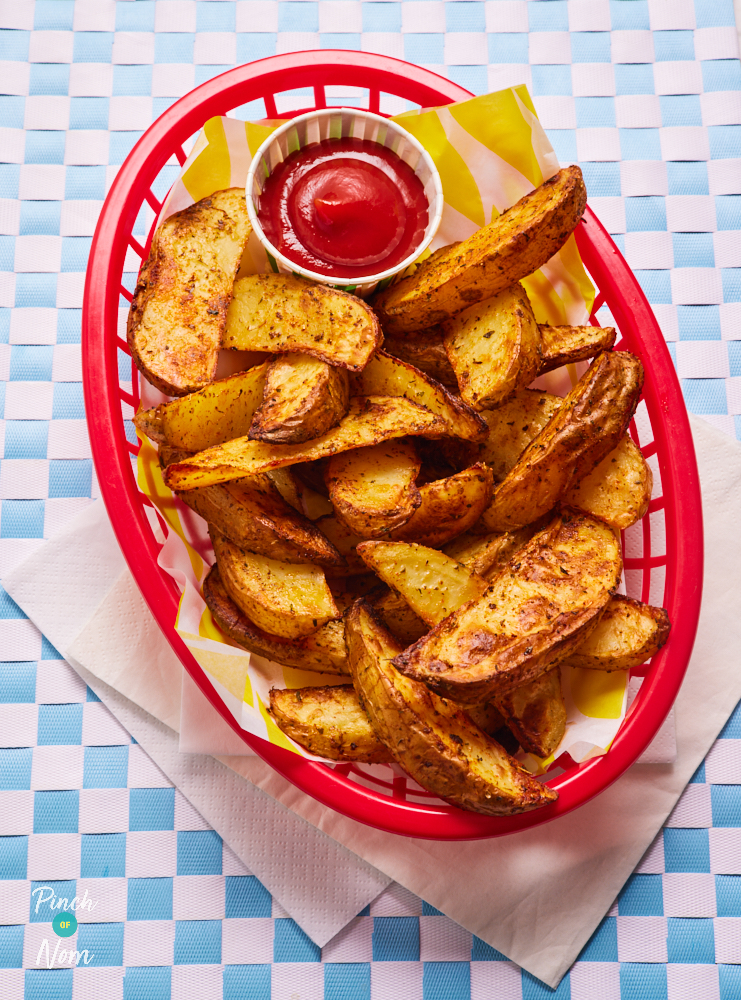 Pinch of Nom's Air Fryer Rustic Potato Wedges served in a restaurant-style basket on colourful baking paper; crispy and golden brown with a pot of ketchup ready for dipping.