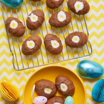 A table is laid with a cheerful yellow and white patterned tablecloth. Pinch of Nom's Chocolate Creme Egg Cakes are set on a cooling rack, with three of them in a yellow bowl nearby. The chocolate cakes are egg-shaped, with white and yellow icing sugar piped into their middles. Easter eggs and decorations are scattered around the table.