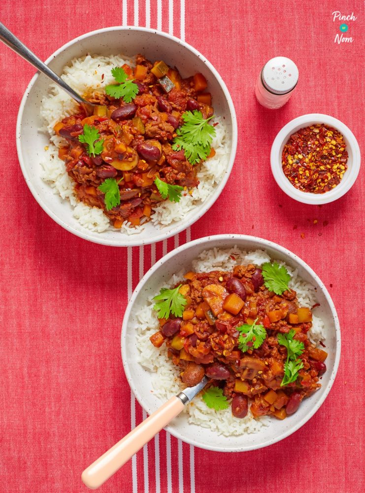 Two bowls of Pinch of Noms Chilli Con Carne are served on a red tabletop. Both portions are topped with fresh coriander leaves; a small pot of chilli flakes waits to one side, waiting to be scattered over.