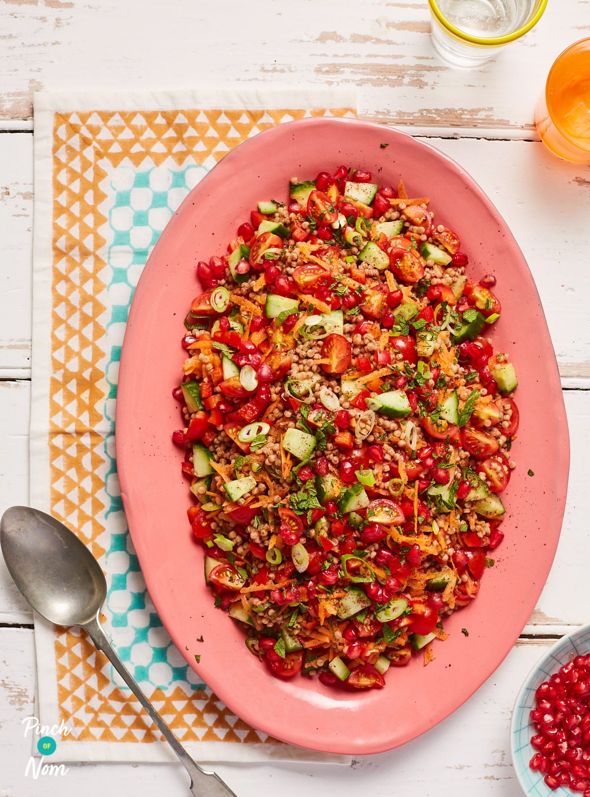 Pinch of Nom's Jewelled Giant Couscous is served on a large bright pink serving dish piled high with plenty of colourful vegetables visibly stirred through.