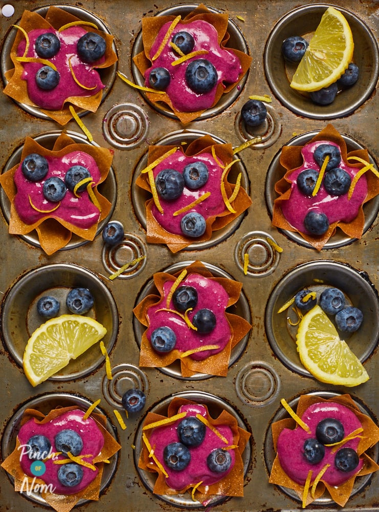 A muffin tray is filled with Pinch of Nom's Lemon and Blueberry Tarts. The tarts are made up of a golden filo pastry case, filled with a vibrant blueberry filling, topped with fresh blueberries and small shavings of lemon zest. A few of the muffin holes are empty, decorated with spare blueberries and lemon.