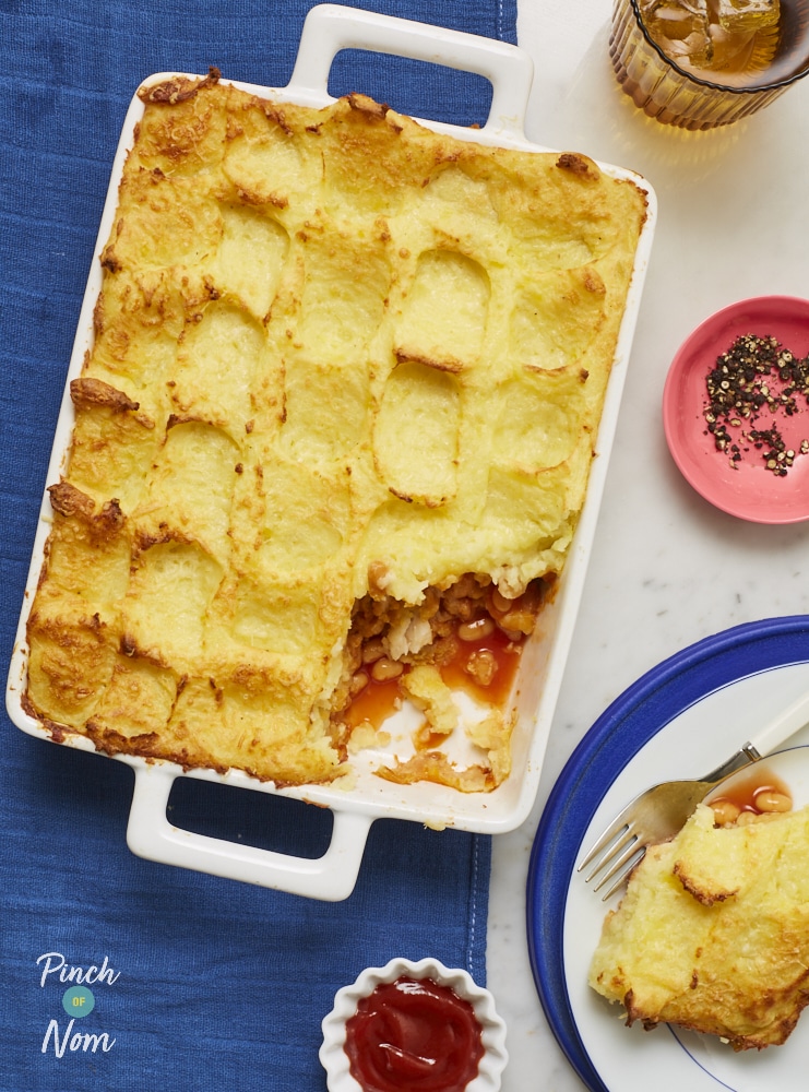 An oven dish of Pinch of Nom's Fish Finger Pie is served. The pie is topped with golden mashed potatoes and a spoonful has been dished onto a dinner plate.
