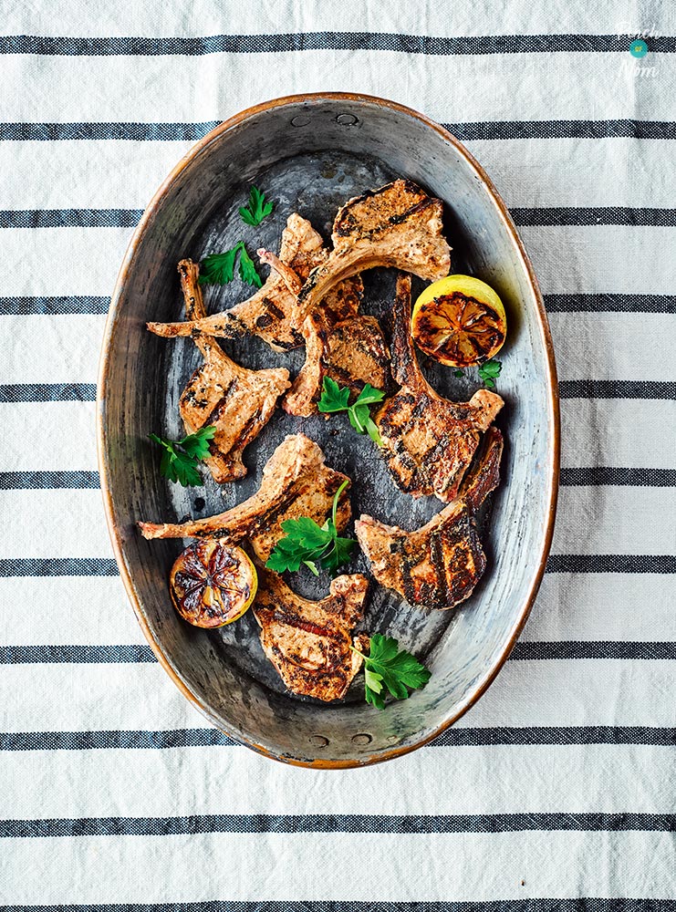 8 of Pinch of Nom's Sumac Lamb Chops are on a large serving plate, on a white, striped tablecloth. Charred lemon wedges are nestled amongst the chops.