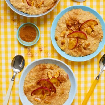 Pinch of Nom's Apple Pie Porridge served in three bowls on a yellow and white striped tabletop, with spoons waiting to dig in. Apple slices and flaked almonds are scattered on top.