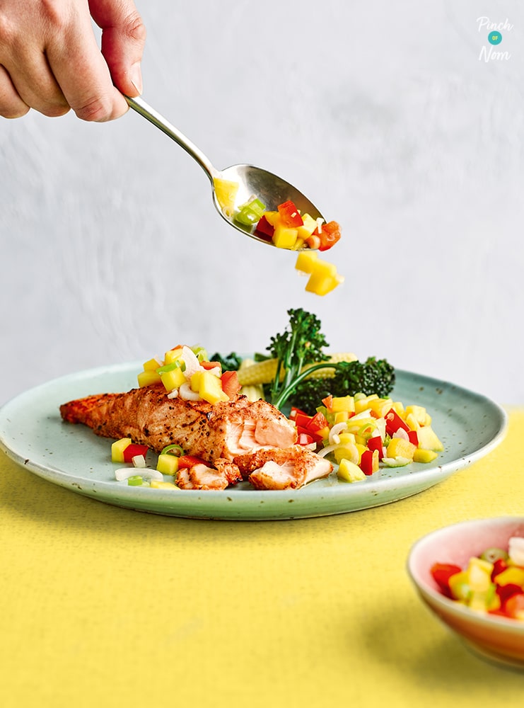 A spoon is drizzling salsa on top of Pinch of Nom's Tandoori Salmon with Mango Salsa. The salmon is plated up with tenderstem broccoli on a yellow tabletop.