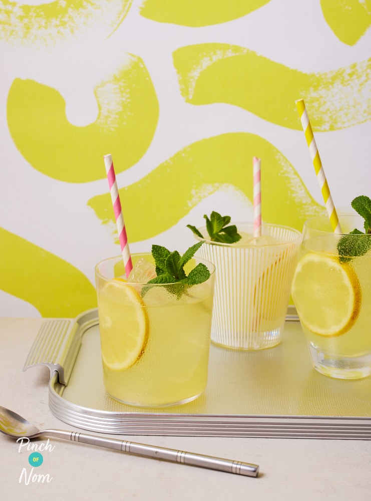 Three glasses are filled with Pinch of Nom's Homemade Lemonade, set on a silver metal tray. Each glass is decorated with a slice fo fresh lemon, a colourful striped straw and a sprig of fresh mint.