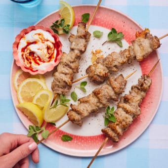 A hand is grabbing a Parmesan Pork Skewer from a red and white plate. The bamboo skewers of pork are coated in golden, melted Parmesan, served on a plate with slices of lemon, scattered herbs and a small bowl of dipping sauce.