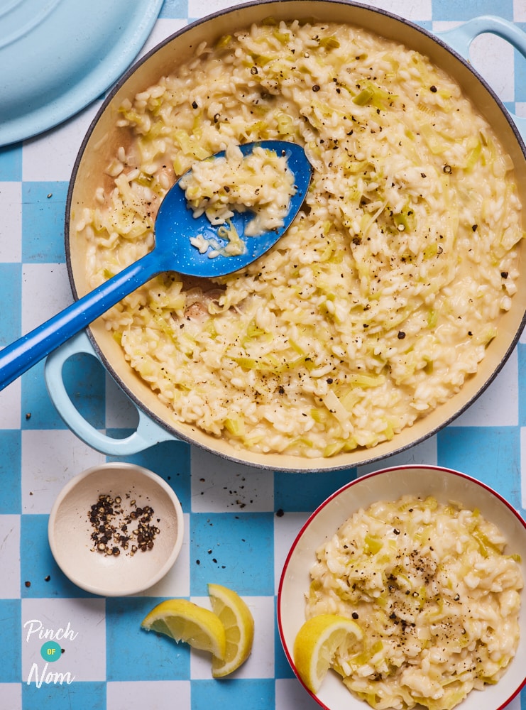 A big pot of Pinch of Nom's Cheesy Leek Risotto is ready to be served. Garnished with fresh lemon and cracked black pepper, a large serving spoon rests in the dish. Nearby, a small bowl has been filled with the low-calorie risotto, ready to eat.