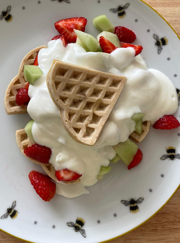 An image of Pinch of Nom's Strawberry and Vanilla Waffles uploaded to the Pinch of Nom website gallery by Louisa. A plate is filled with heart-shaped waffles, chopped fruits and sweetened yoghurt.