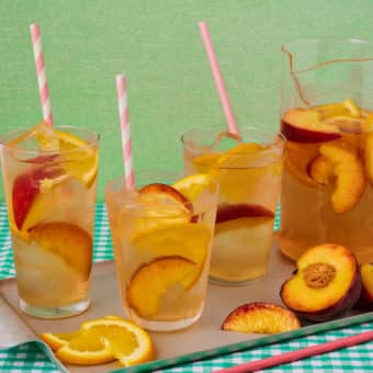 Pinch of Nom's Pink Sangria is served with lots of sliced peaches and oranges, in 4 glasses with pink and white striped straws. Peach and orange slices are also scattered on the board around the glasses, which rest on a gingham tabletop.