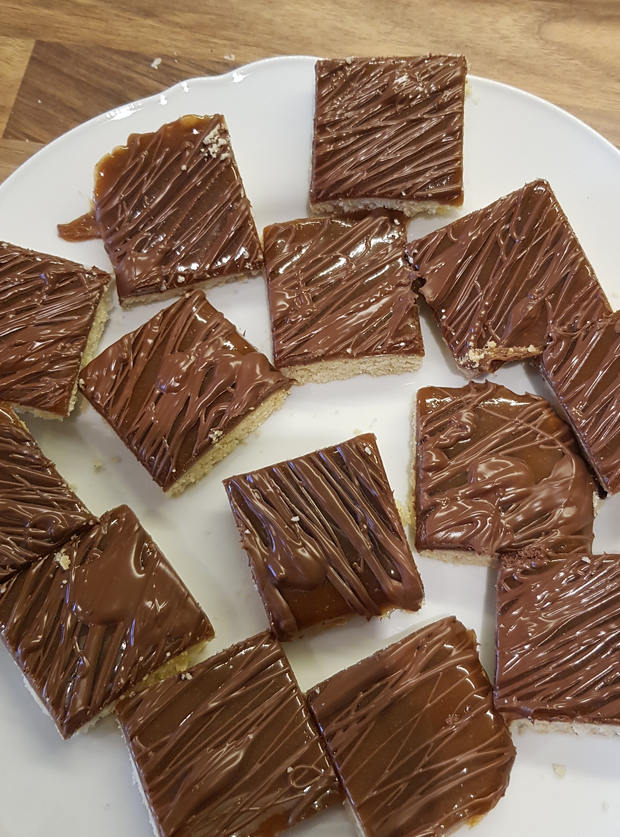 An image of Pinch of Nom's Millionaire's Shortbread recipe made at home by Zoe, uploaded to Pinch of Nom's website gallery. A plate is covered with shortbread pieces covered in a layer of gooey caramel and a chocolate drizzle.