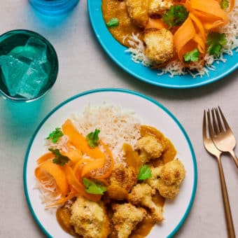 Two plates of Pinch of Nom's Caulifower Katsu sit on a kitchen counter, next to forks and glasses of ice water. The katsu curry has been served with a portion of fluffy rice and ribbons of crunchy carrot.
