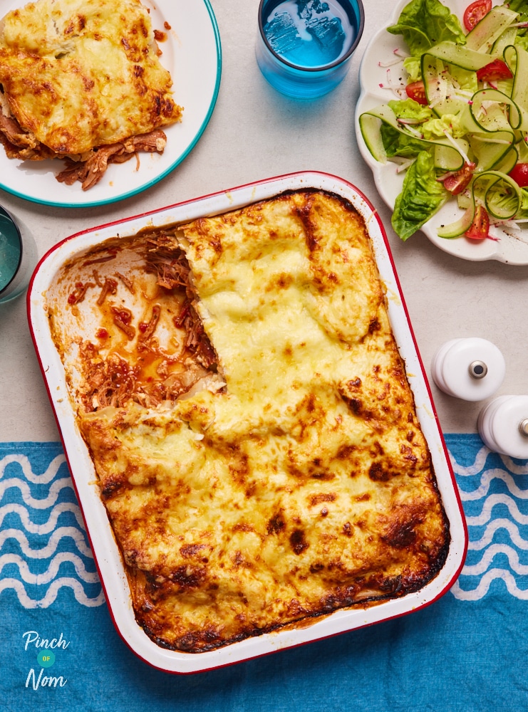 Pinch of Nom's Pulled Pork Lasagne is in the middle of the table, served in a large rectangular oven dish. A corner has been served onto a small plate nearby, and a side salad is waiting on a separate plate. The lasagne is golden brown and cheesy, with a BBQ pork filling. 