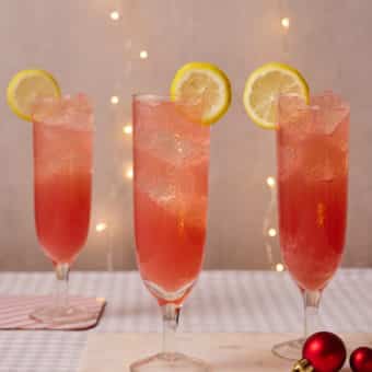 Pinch of Nom's Cranberry Fizz is served in three flutes on a table decorated with festive lights and sparkly baubles. Each glass has a lemon wedges onto the rim for a touch of luxury.