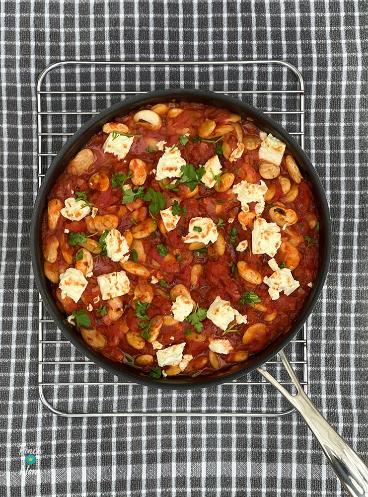 A shallow pan is filled with Pinch of Nom's Greek-Style Baked Beans. Butter beans are nestled in a rich, tomato sauce, with crumbled feta cheese and fresh herbs scattered on the top.
