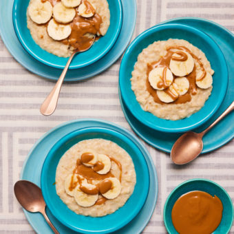 Three bowls of Pinch of Nom's Biscoff and Banana Porridge are served drizzled with melted Biscoff spread, topped with freshly-sliced bananas. A side bowl of melted Biscoff spread is on the side, with spoons waiting to tuck in.
