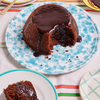 A table is set with a white and multi-coloured striped tablecloth. In the centre, a light blue patterned plate is filled with a dome-shaped chocolate cake, with chocolate sauce poured over the top and running down the sides. A slice has been cut from the front of the cake and served on a small white plate that can be seen in the bottom left of the photo. This is Pinch of Nom's slimming-friendly Chocolate Sponge Pudding.
