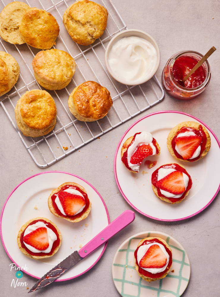 Pinch of Nom's Cream Tea Scones are served on an oven tray; some are sliced in half and plated up with layers of jam, cream and freshly-sliced strawberry on top.