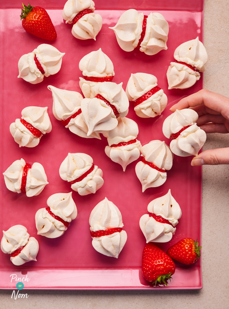A bright pink tray is filled with Pinch of Nom's Creamy Strawberry Mini Meringues. On the right hand side, a hand is lifting on of the meringues from the tray. Each one is made up of two meringue kisses, sandwiched together with a strawberry filling.
