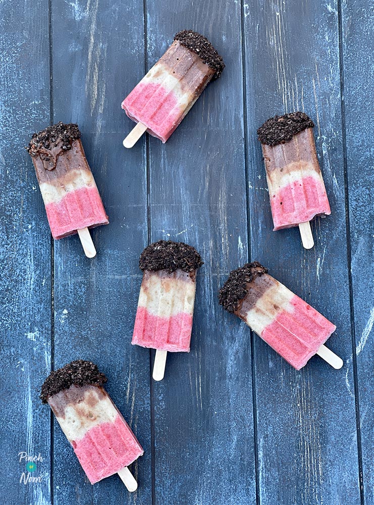 6 of Pinch of Nom's Neapolitan Ice Pops from the Budget cookbook are arranged on a wooden tabletop with a drizzle of melted chocolate and crushed biscuits on the top layer.
