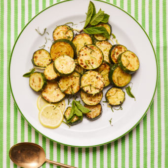 A table is covered with a green and white striped tablecloth. A large white plate with a green rim is filled with Pinch of Nom's Lemon and Mint Courgettes. A large serving spoon is nearby. The golden brown courgettes are sliced into round discs and garnished with fresh mint and lemon slices.