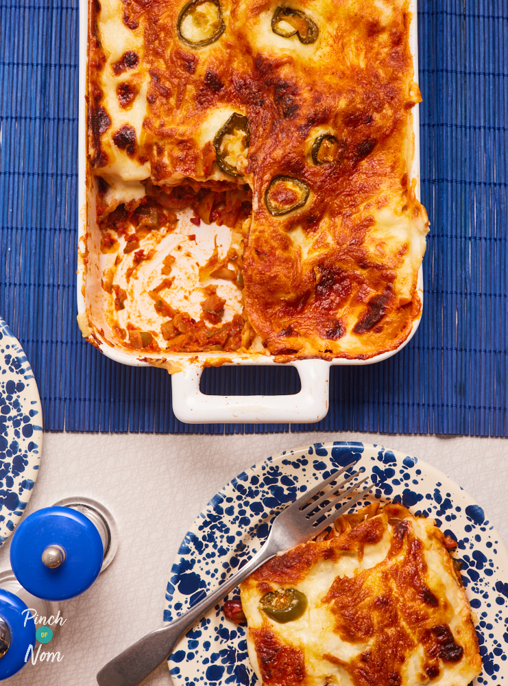 Pinch of Nom's Chilli Bean Lasagne is served fresh from the oven in a large casserole dish. The cheesy top is melty and golden brown, with jalapeños poking through. A single portion of lasagne waits on a plate to the side for someone to tuck in.