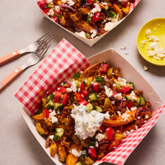 Takeaway style food cartons are filled with Pinch of Nom's Loaded Lamb Sweet Potato Fries. The golden fries are topped with lamb mince, chopped vegetables and crumbled feta cheese.