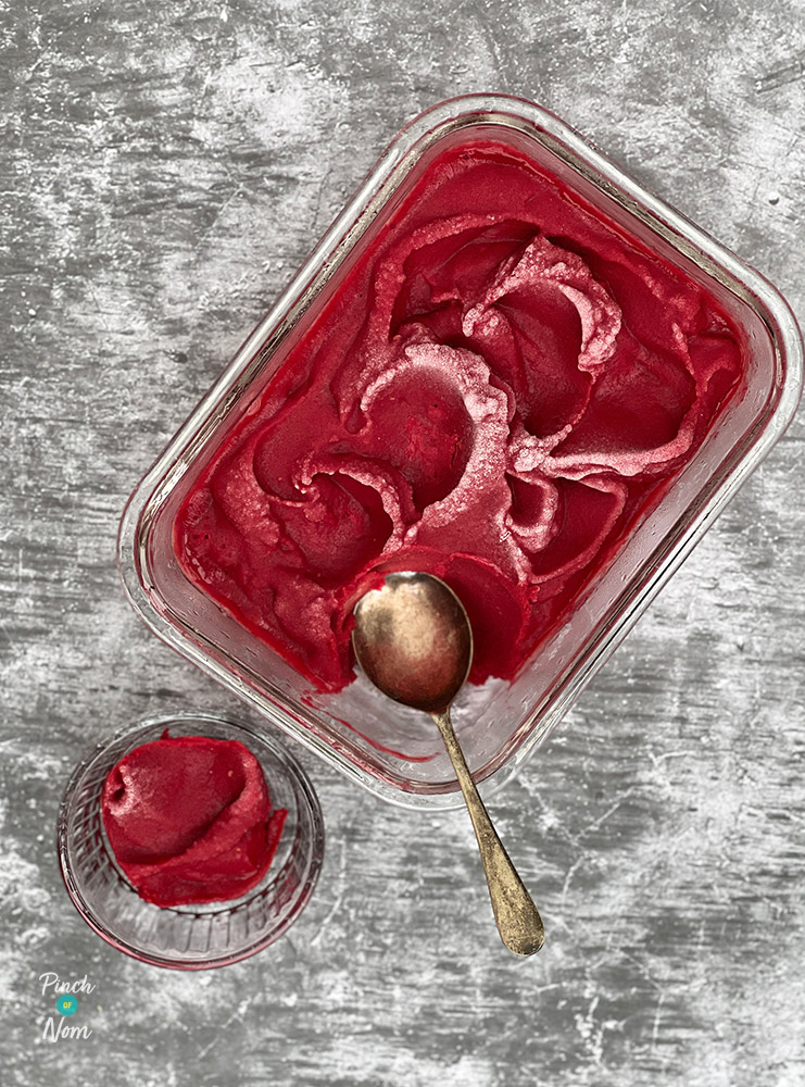 Pinch of Nom's Raspberry Sorbet is on a tabletop fresh from the freezer in a glass, freezer-proof dish; an spoon is scooping a serving into a small glass ramekin.