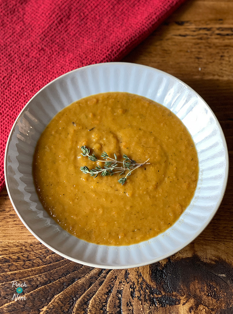 A white bowl is filled with Pinch of Nom's Roasted Root Vegetable Soup. The soup is thick and creamy, with a deep orange colour and a simple garnish of fresh herbs.