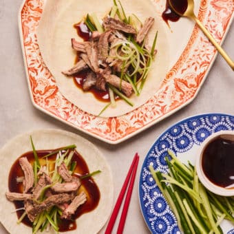 A table is laid with chopsticks, a serving platter of crispy duck, shredded cucumber, spring onions, pancakes and hoisin sauce. Two pancakes are loaded up with fillings, ready to be rolled up and eaten.